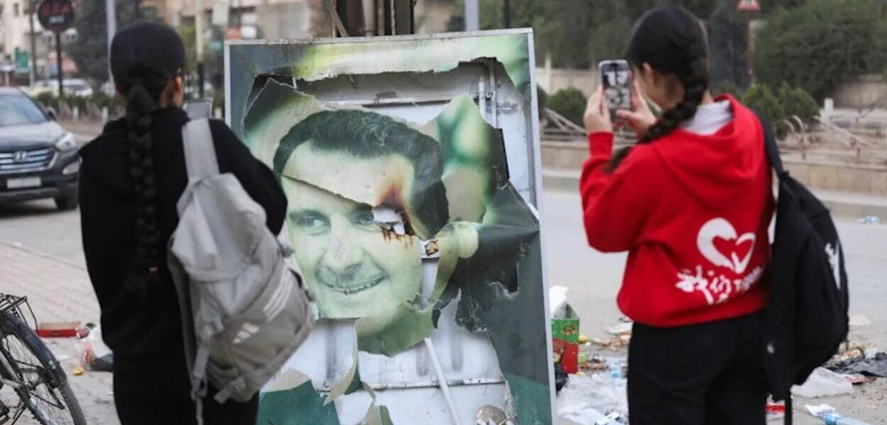 Mulheres tiram fotos de uma imagem danificada do presidente sírio Bashar al-Assad