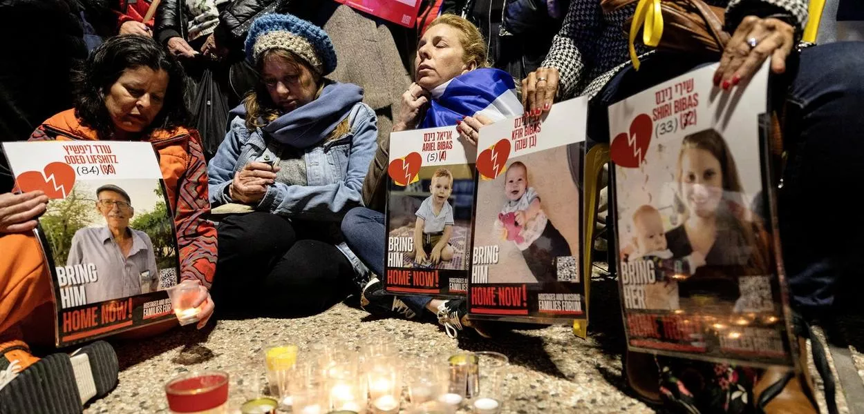 Israelenses sentam-se juntos enquanto acendem velas e seguram cartazes com as imagens de Oded Lifschitz, Shiri Bibas e seus dois filhos, Kfir e Ariel Bibas, capturados em 7 de outubro de 2023
