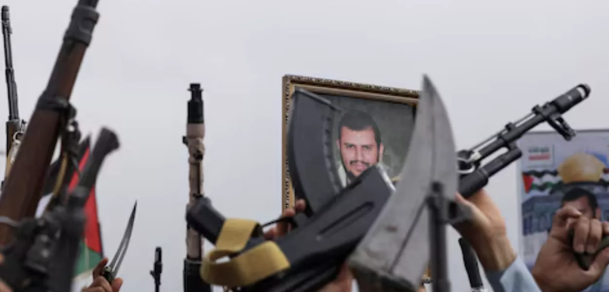 Um cartaz do líder Houthi, Abdul Malik al-Houthi, é segurado por um manifestante enquanto outros seguram suas armas durante uma manifestação em apoio aos palestinos na Faixa de Gaza, em Sanaa, Iêmen, em 26 de julho de 2024