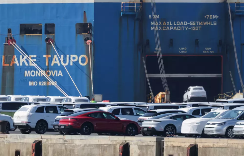 Um carro do Grupo Volkswagen é carregado em um navio de carga enquanto carros de luxo Porsche e outros VW destinados à exportação para os Estados Unidos aguardam para serem carregados no porto de Emden, perto do estuário, onde o Rio Ems deságua no Mar do Norte, em Emden, Alemanha, em 2 de abril de 2025