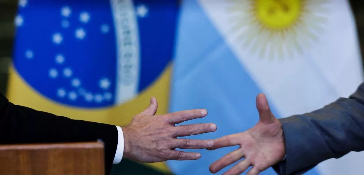 Aperto de mãos em frente às bandeiras do Brasil e da Argentina