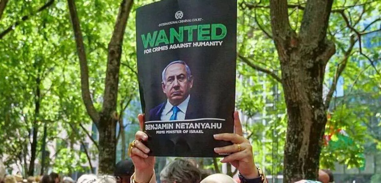 Uma mulher segura um cartaz onde se lê "Procurado por crimes contra a humanidade, Benjamin Netanyahu", durante a manifestação "Red Line For Gaza", em 15 de junho de 2025, em Bruxelas, Bélgica.