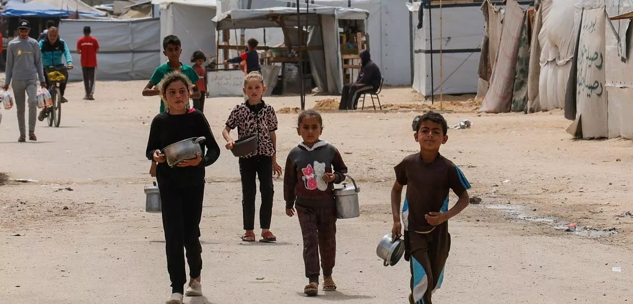 Crianças palestinas caminham enquanto pessoas esperam para receber comida preparada por uma cozinha comunitária, em Khan Younis, no sul da Faixa de Gaza