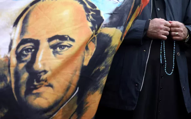 Um padre segura um rosário ao lado de uma bandeira com o retrato do falecido ditador espanhol Francisco Franco, do lado de fora do cemitério Mingorrubio-El Pardo, em Madri, Espanha, em 24 de outubro de 2019
