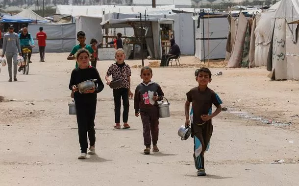 Crianças palestinas caminham enquanto pessoas esperam para receber comida preparada por uma cozinha comunitária, em Khan Younis, no sul da Faixa de Gaza - 06/04/2025
