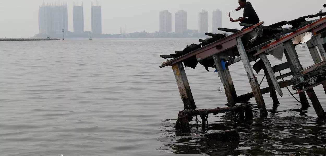 Um jovem sobre os destroços de um barco de madeira, durante uma atividade de pescaria, enquanto a névoa de poluição cobre os céus de Jacarta, na Indonésia, em 26 de julho de 2018